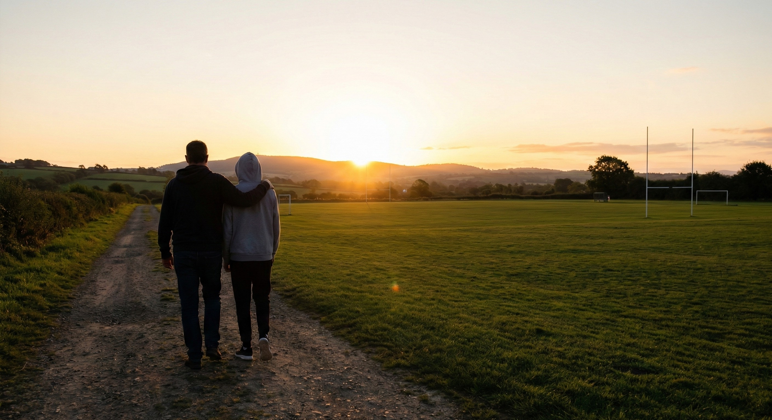 Parent walking with their arm around a teen athlete near a sports field at sunset. Depicting unconditional support and connection over performance for families navigating youth sports pressure.