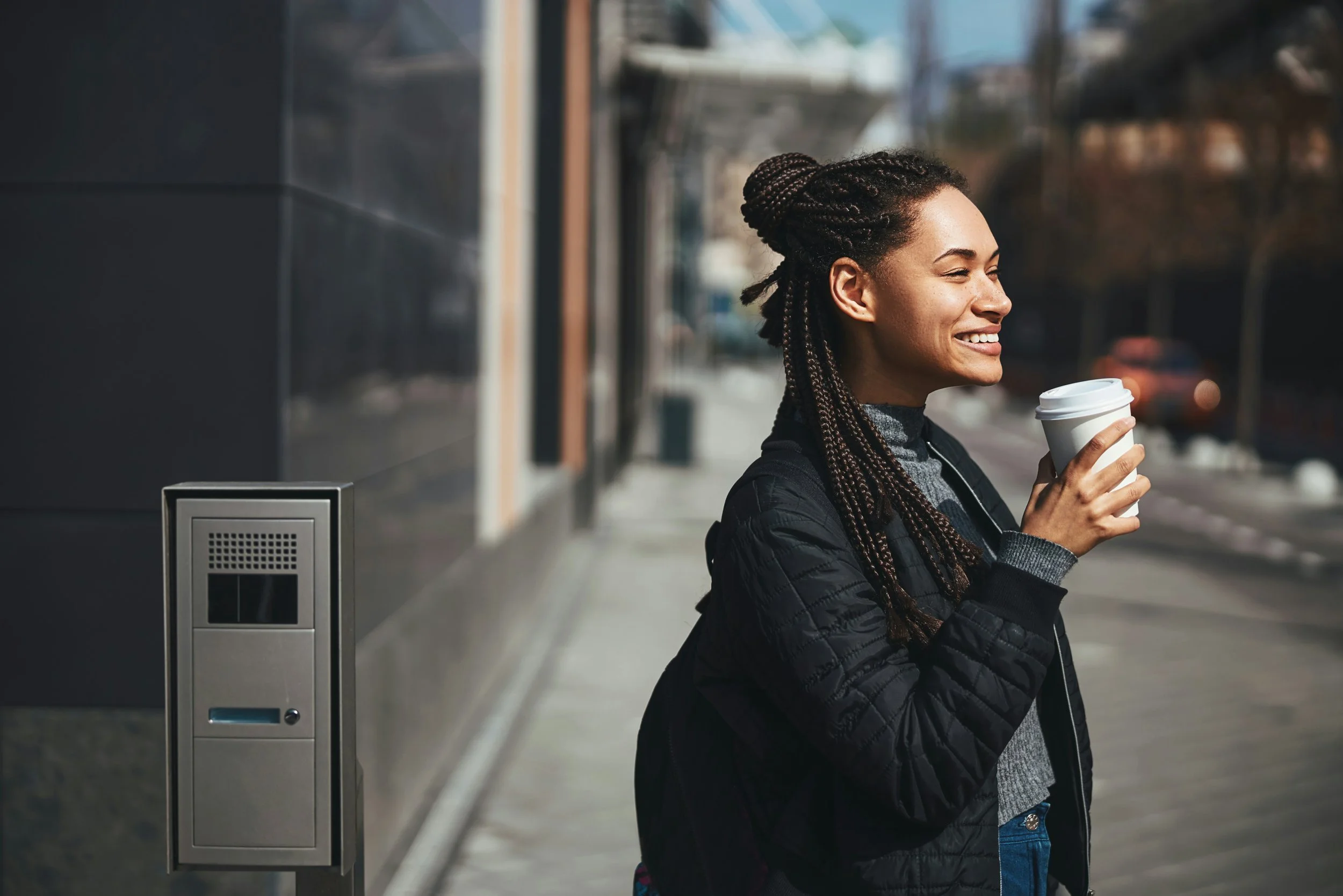 Smiling young woman holding coffee on a sunny city sidewalk, feeling lighter after working through burnout in therapy