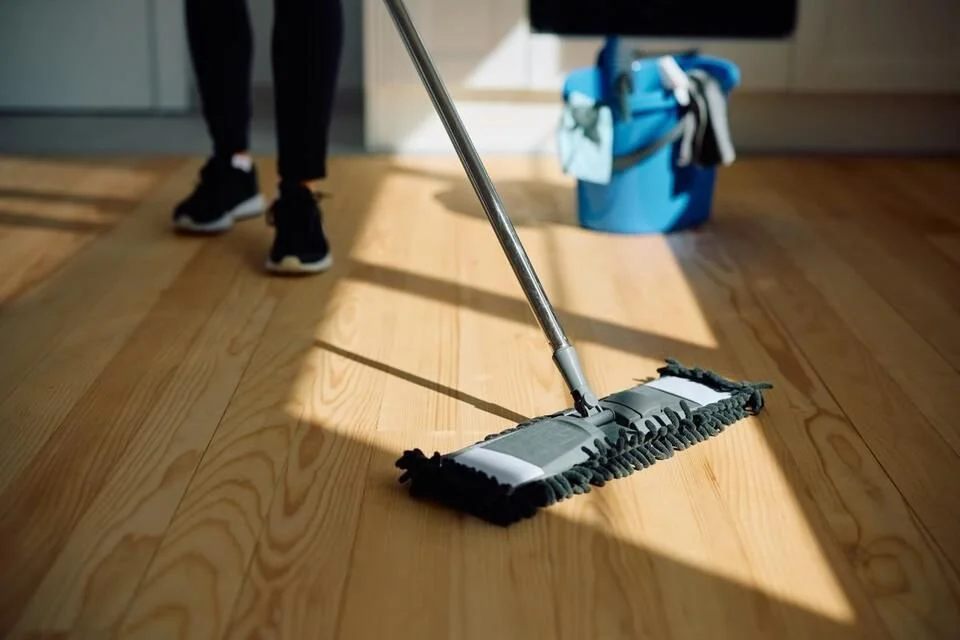 Person som använder en mopps för att torka golvet i ett rum med trägolv, med en blå hink i bakgrunden.