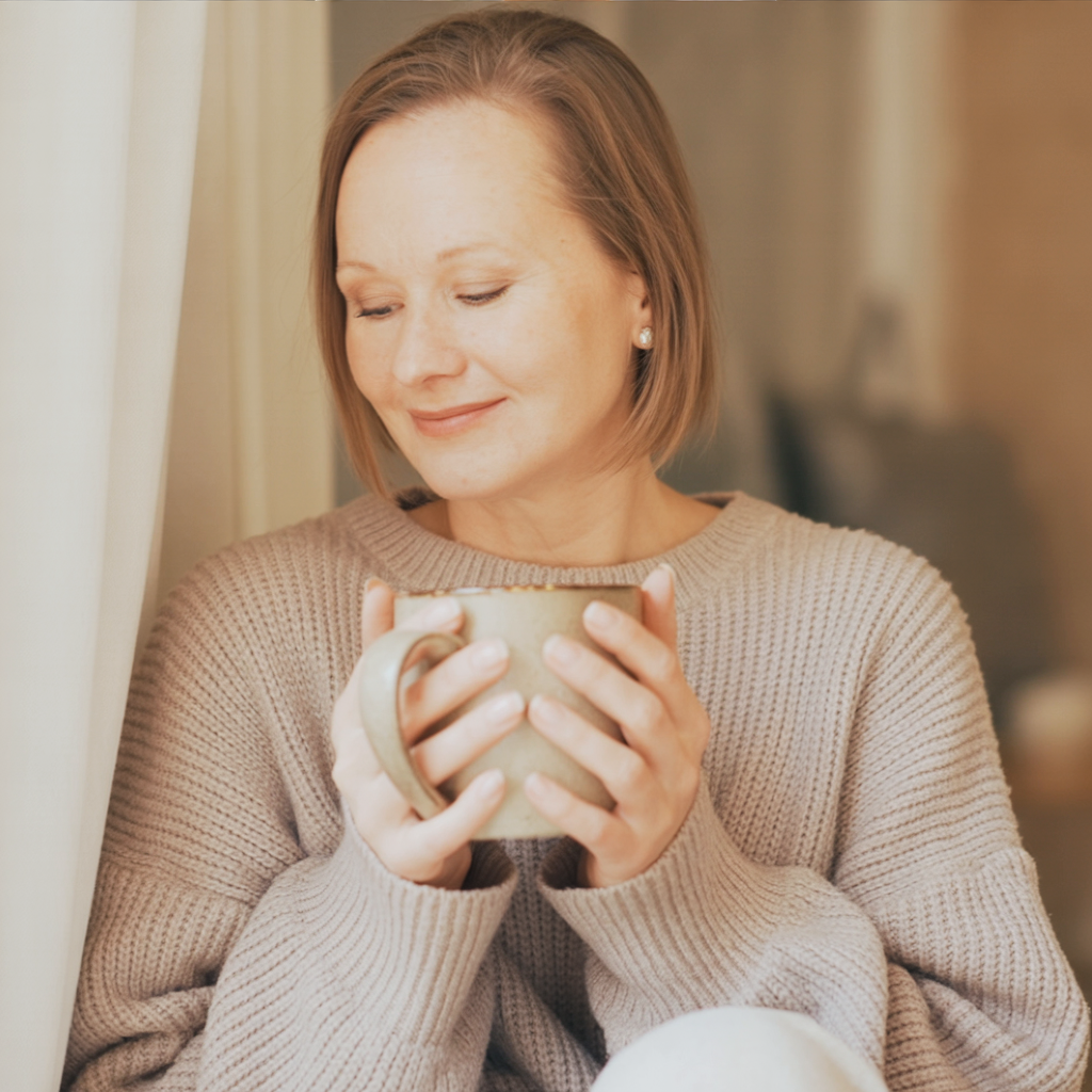 A woman wrapped in a soft knit sweater, sitting by a window with morning light, hands around a warm mug