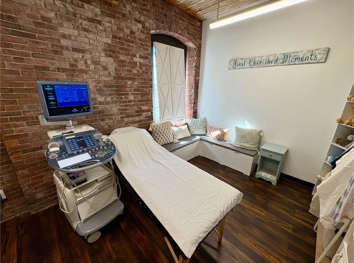 Warm and welcoming elective ultrasound studio with an exam bed, professional ultrasound equipment, exposed brick walls, and family seating area.