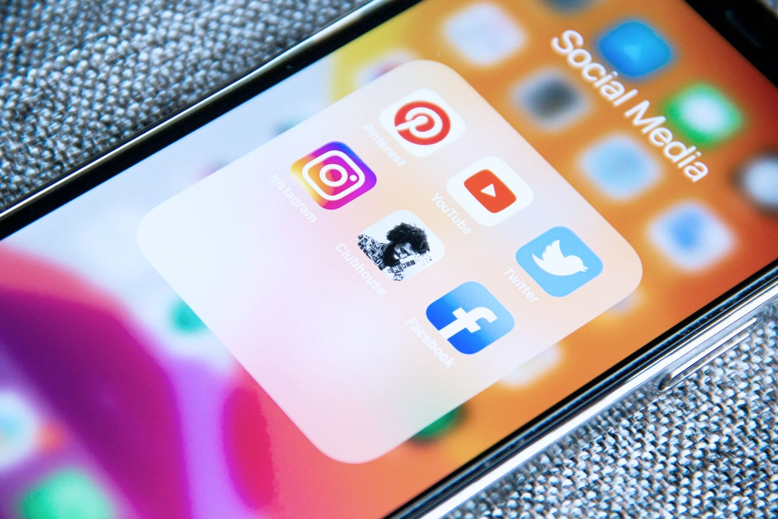 Close-up of a smartphone screen displaying social media app icons including Pinterest, YouTube, Instagram, Twitter, Facebook, and Clubhouse within a folder labeled 'Social Media' placed on a gray textured surface.