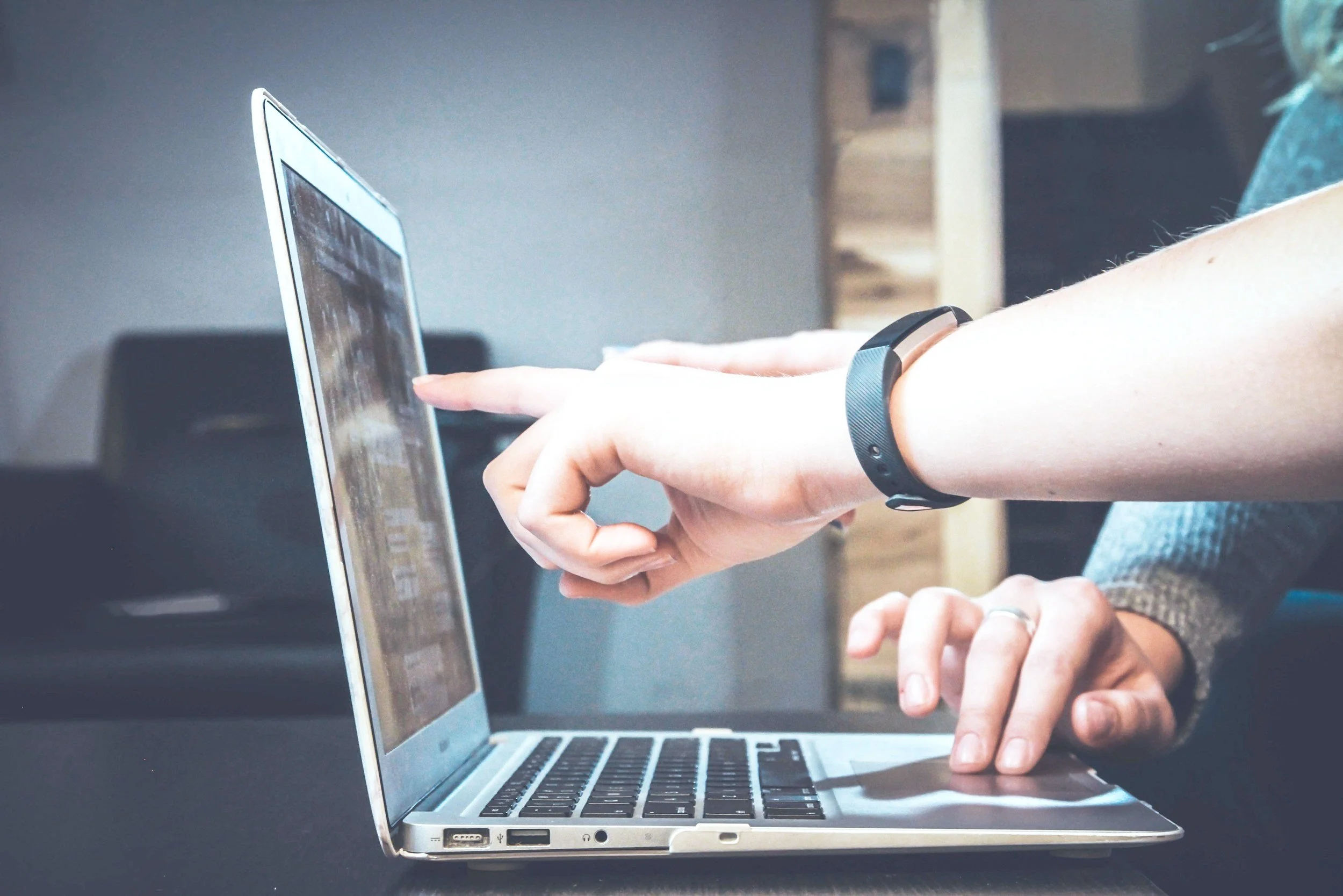 Person using a laptop with a black wristband on their right wrist, pointing at the screen with their right index finger, while their left hand rests on the keyboard.