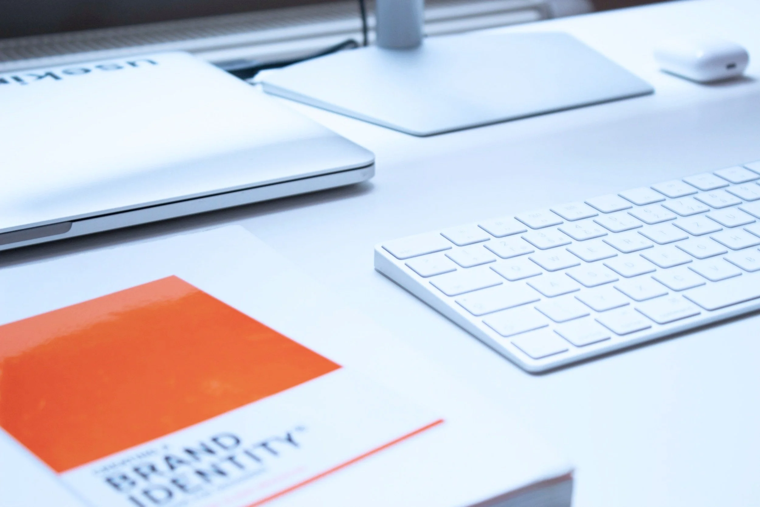 A workspace with a closed MacBook, an Apple Mouse, a white keyboard, a white mouse pad, and a book titled "French Identity" on a white desk.
