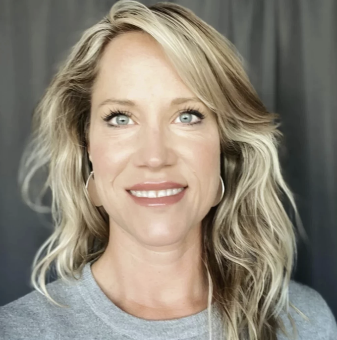 A woman with shoulder-length blonde hair and blue eyes smiling at the camera, wearing a gray top and earrings, with a dark background.