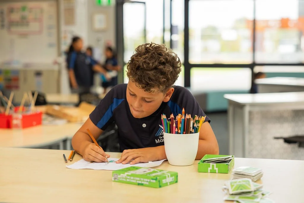A young boy with curly hair is sitting at a school desk, coloring with colored pencils on paper.