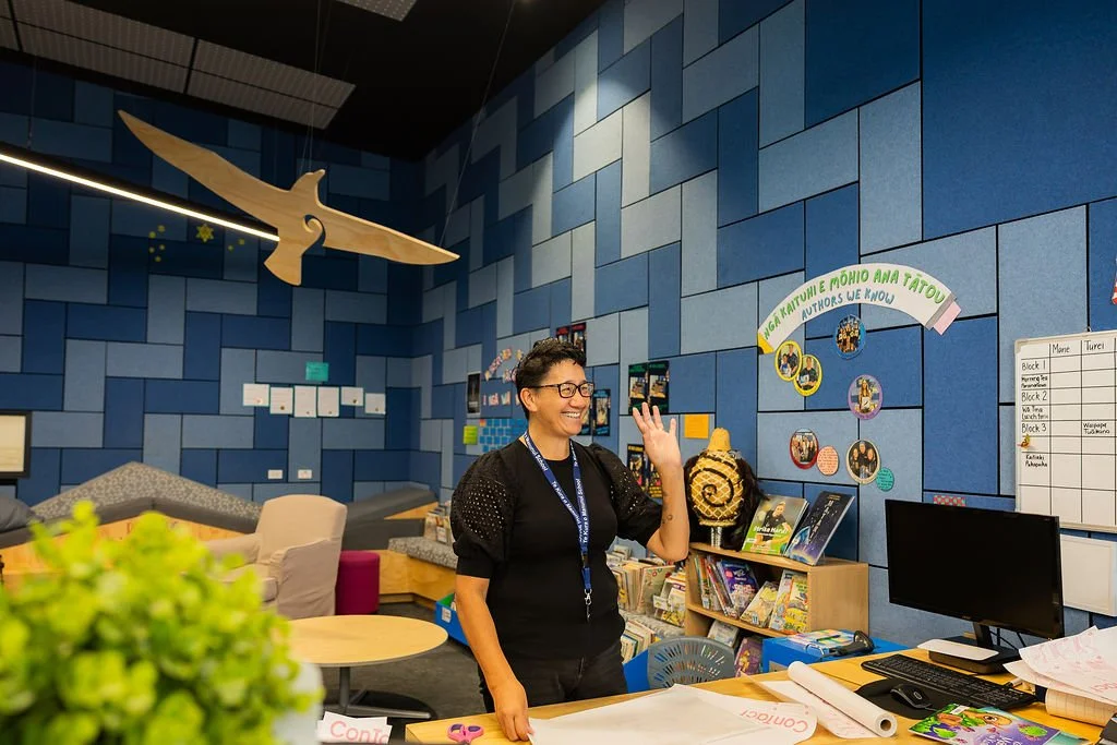A woman with short dark hair and glasses smiling and waving in a colorful school library.