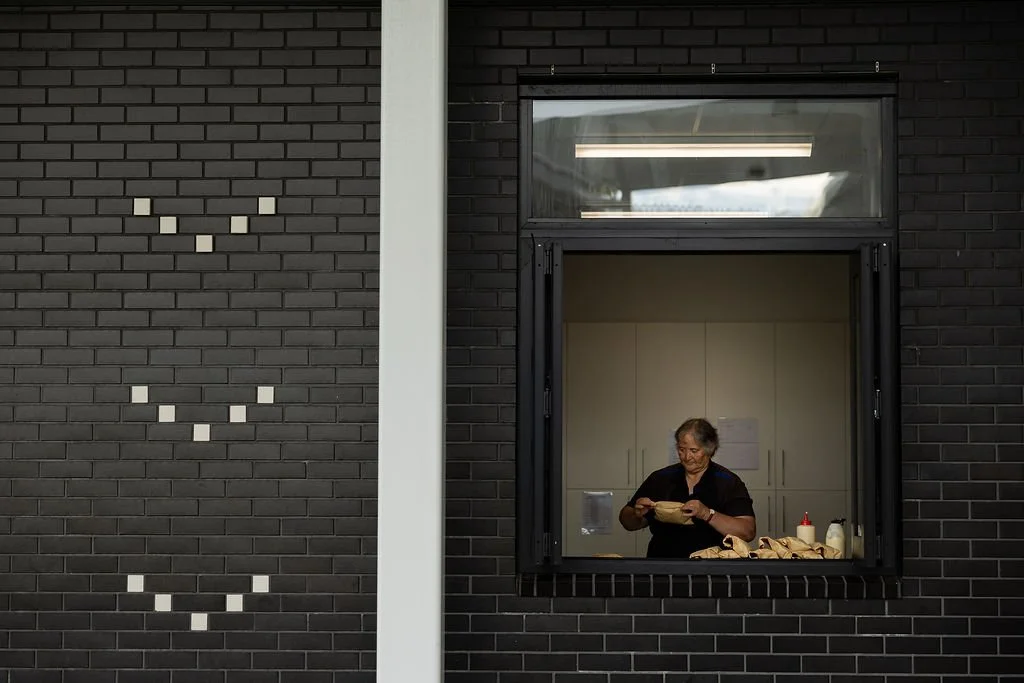 A woman working in a kitchen seen through a large window in a black brick wall