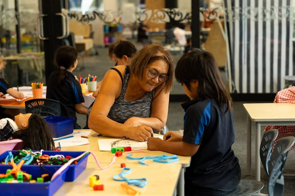 A kaiako and a young girl sit at a desk in a classroom, working on a project together, surrounded by art supplies and toys.