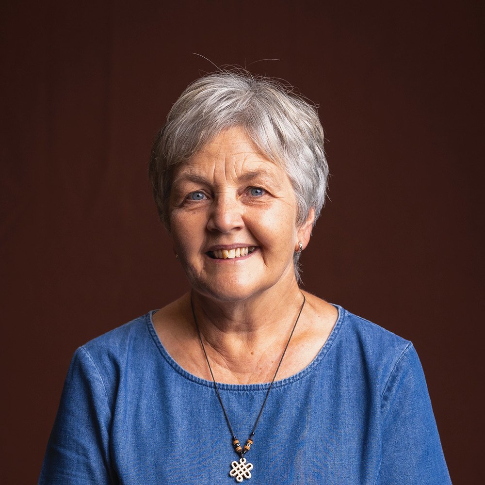 Smiling woman with short brown hair wearing a light green shirt and necklace, against a brown background.