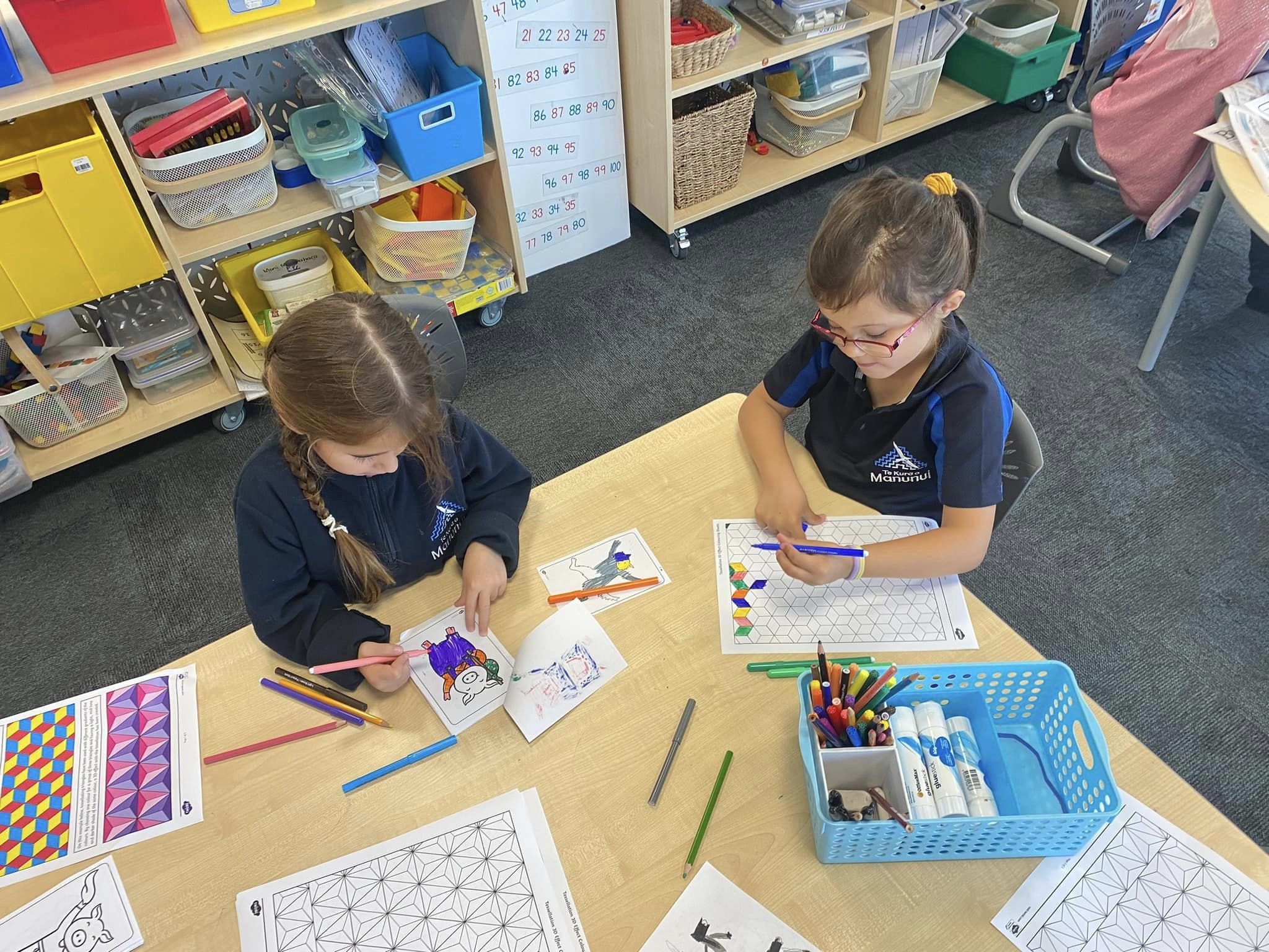 Two children sitting at a table in a classroom, engaged in coloring activities with markers and pencils. The table is covered with sheets of geometric patterns and coloring pages. A blue basket holds art supplies. Shelves in the background contain va