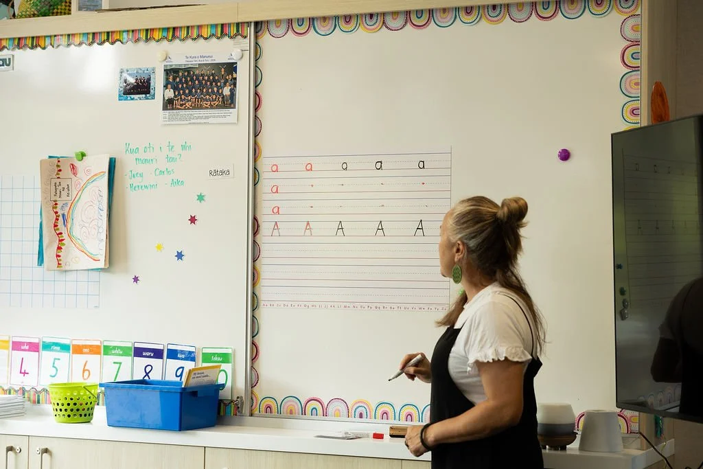 A teacher writing on a whiteboard in a classroom, with educational decorations and a photo collage on the wall.