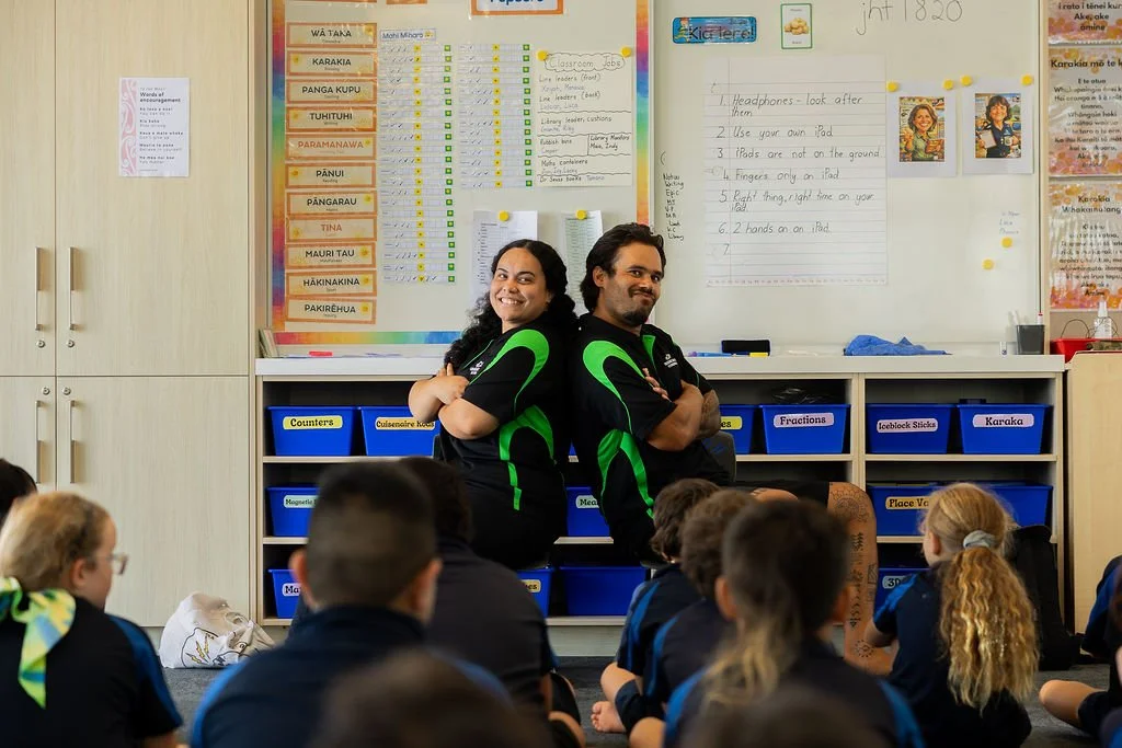 Kids Can assistants in front of a classroom of young students. The students are sitting on the floor, facing the front, wearing school uniforms. The background has educational posters, charts, and a whiteboard.