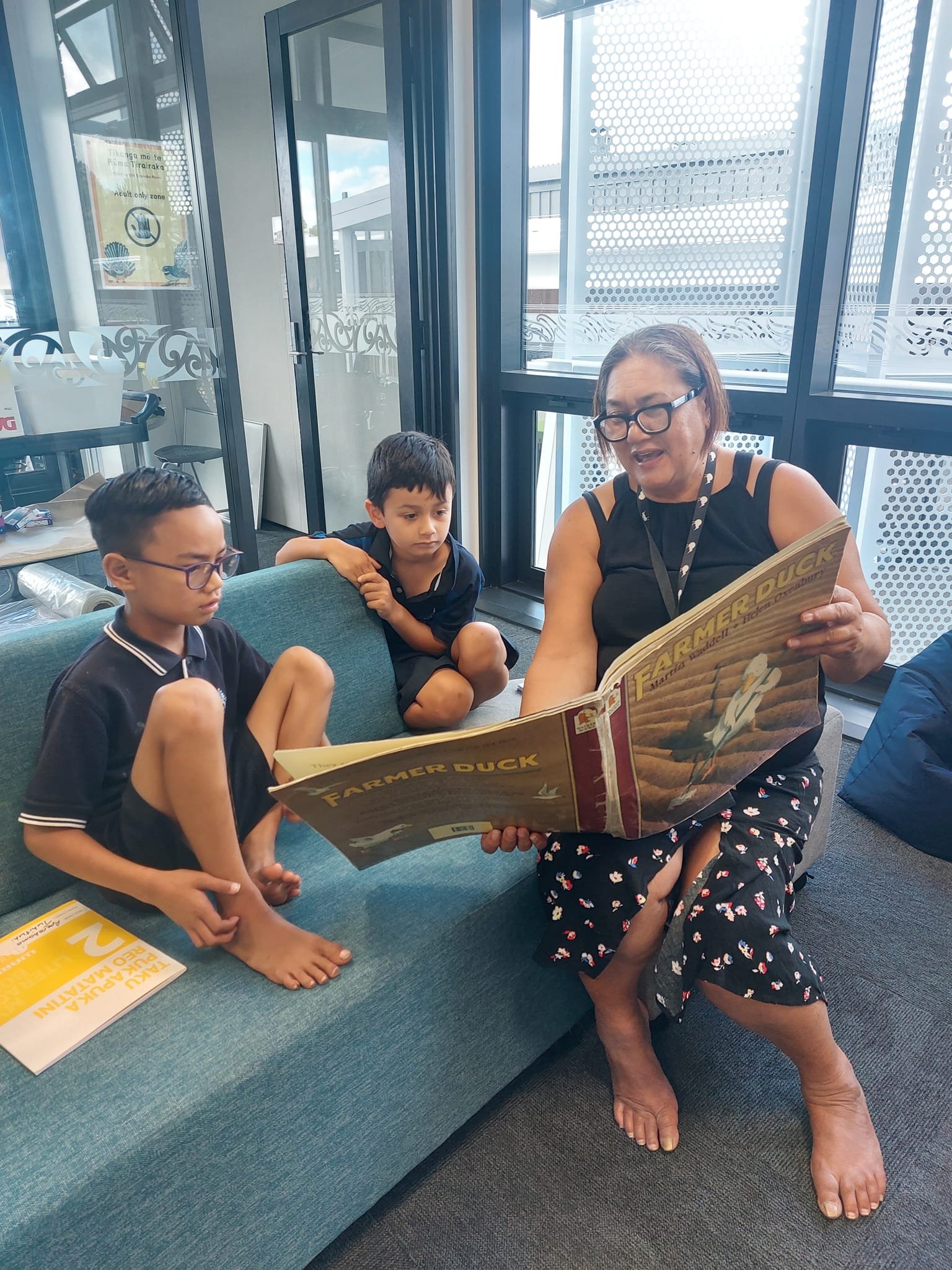 A woman and two young boys sitting on a sofa in a library setting. The woman is reading a book titled 'Farmer Duck' to the children. They appear to be engaged in a storytelling session. The room has large glass windows and a modern interior.