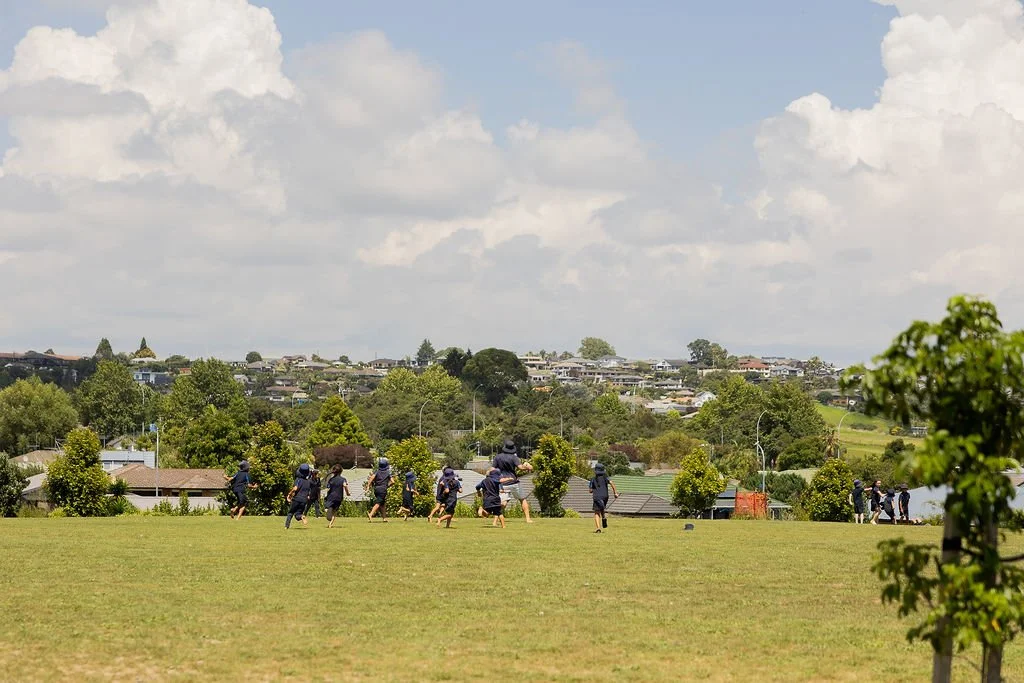 Group of children or students walking across a grassy field in a park or schoolyard, with trees and a hill with houses in the background under a partly cloudy sky.
