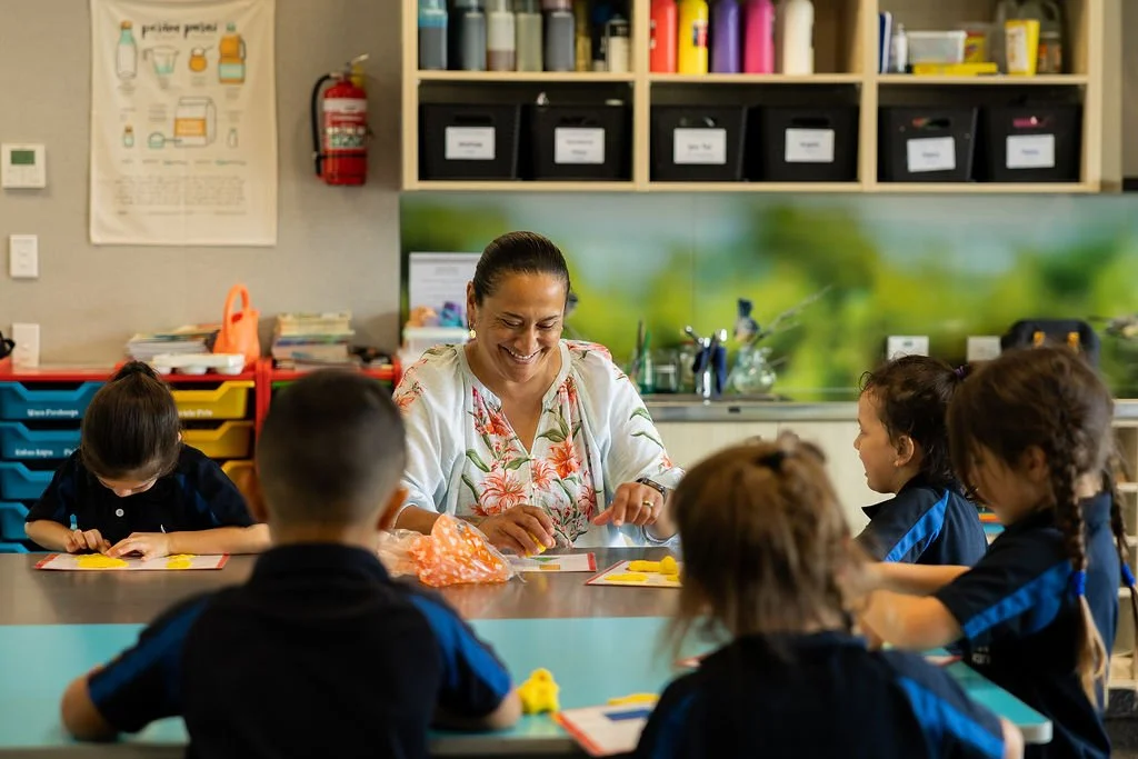 A teacher sitting at a table with five young students in a classroom, engaging in a craft activity. The children are wearing matching black and blue uniforms. The classroom has educational posters, storage bins, and a colorful background.