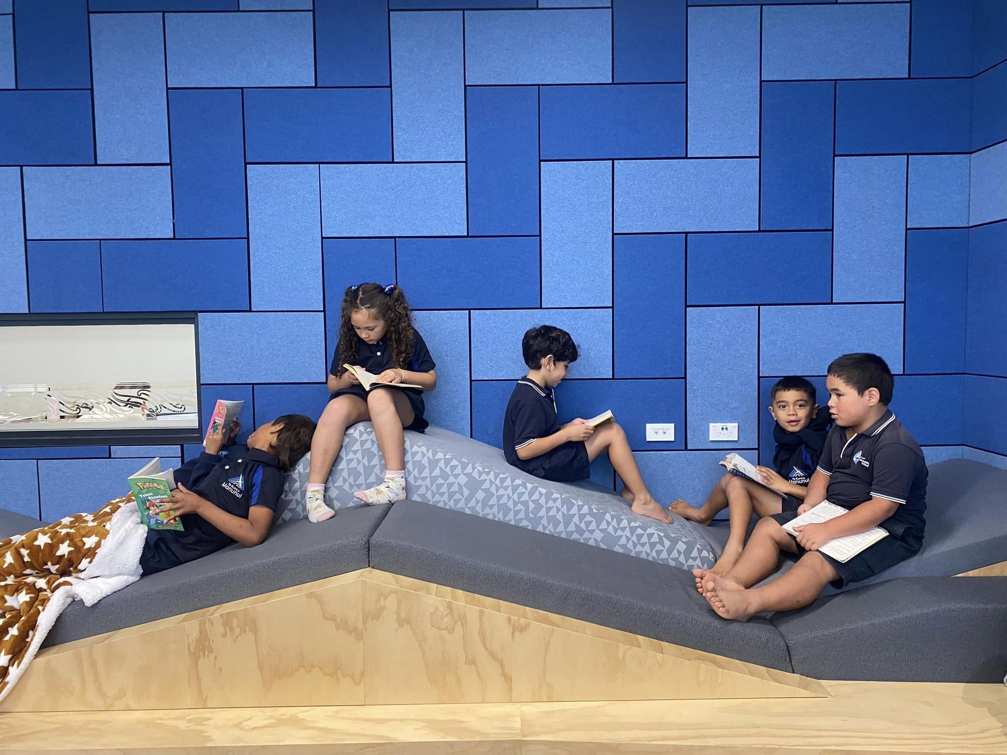 Children sitting and reading on gray cushions against a blue tiled wall.