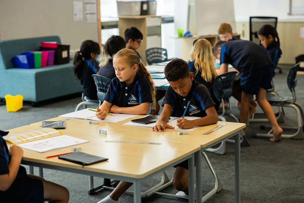Students working at desks in a classroom, some writing and others collaborating.