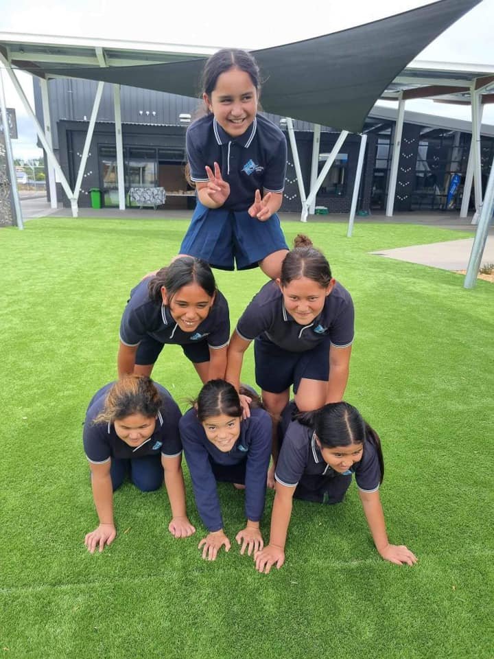 Group of six kids in navy uniforms forming a human pyramid on a grassy field with a modern building in the background.