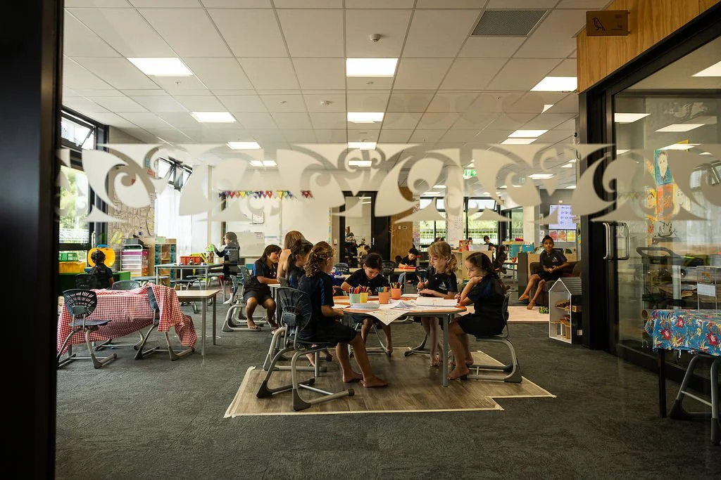 Children sitting at tables in a classroom or activity room, engaged in arts and crafts, with adult supervisors in the background.