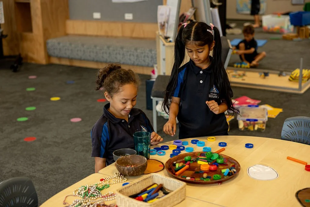 Two young girls, wearing navy blue uniforms, are playing with colorful educational toys and game pieces at a round table in a classroom. Background shows a play area with children and various toys.