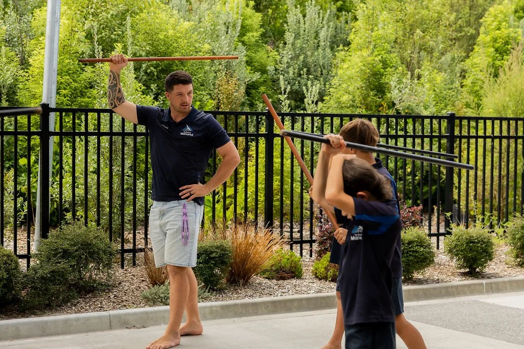 A man in a navy shirt and shorts instructing two children in mau rākau with wooden sticks outdoors, surrounded by a black fence and greenery.