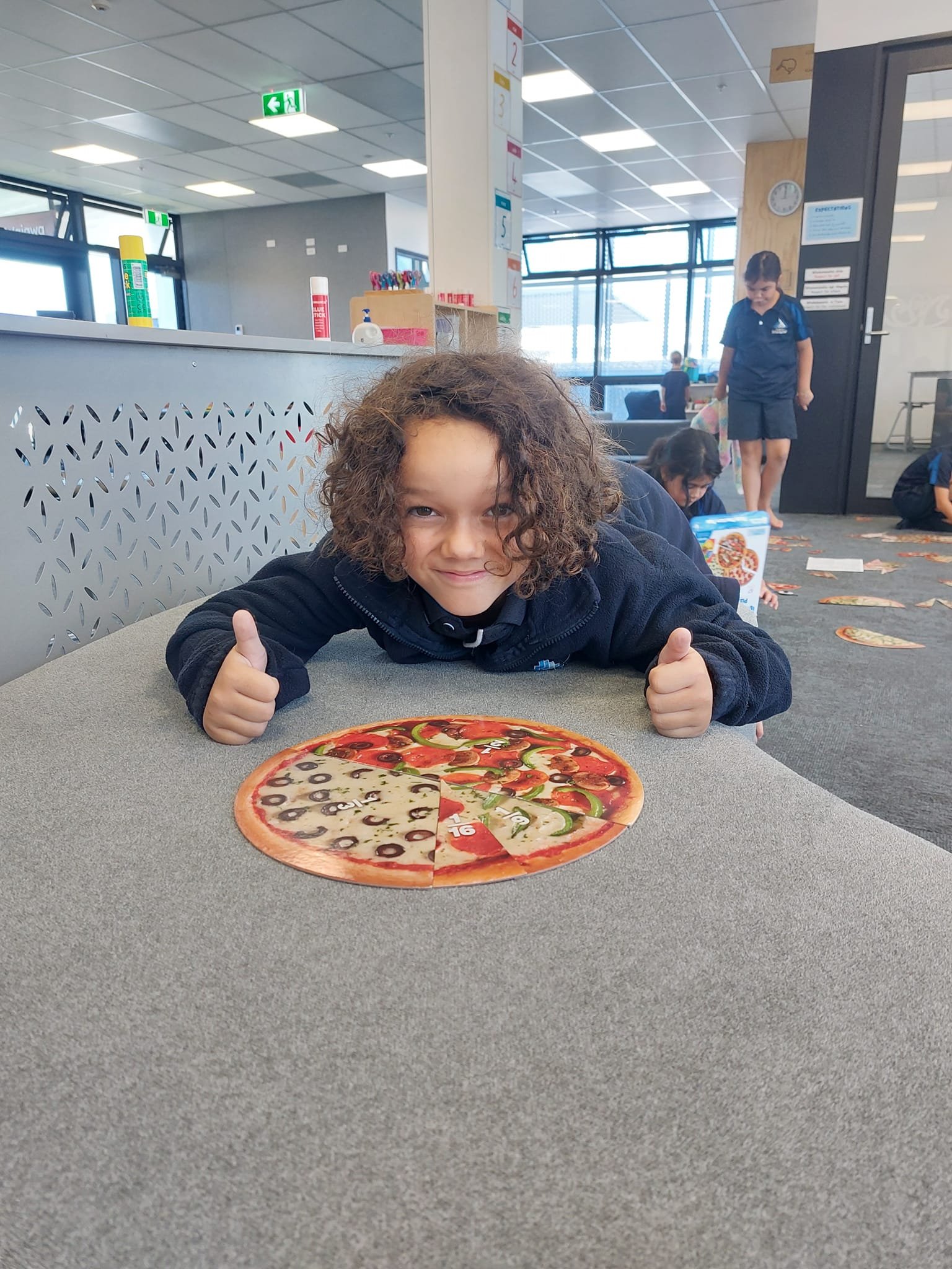 Child giving thumbs up near a pizza puzzle on a table with other children in the background in a classroom setting.