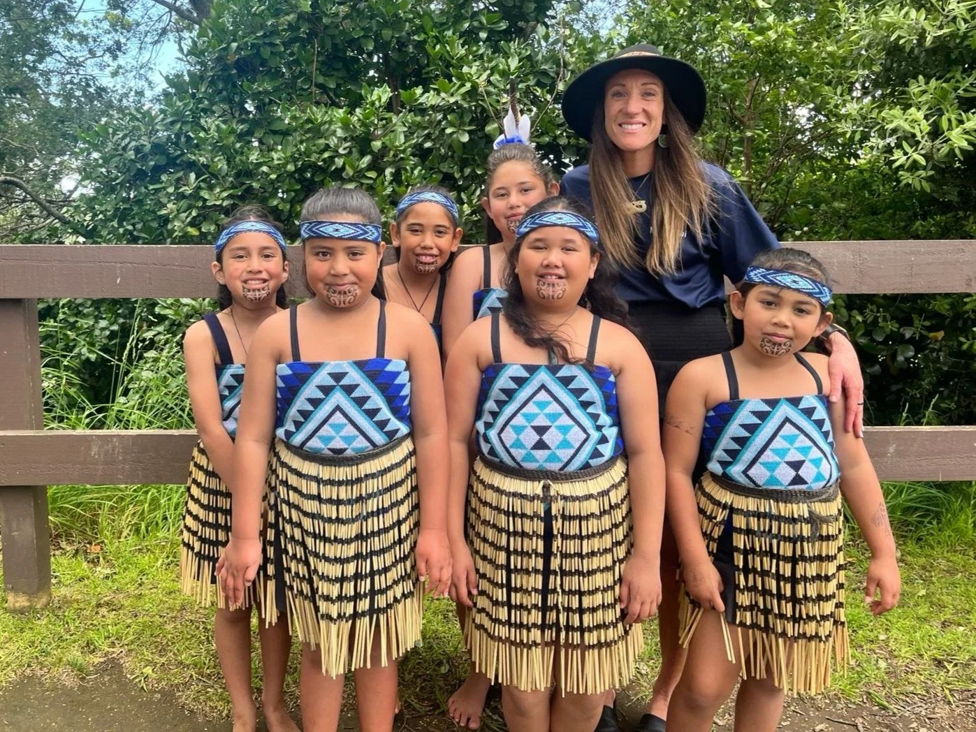 Group of six young girls in traditional Māori kapa haka gear with blue and black geometric patterns, posing with a woman in a black hat and navy blue shirt outdoors next to greenery.