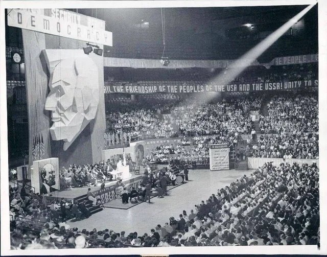 Vintage black and white photo of a large indoor gathering with a stage decorated with a large face sculpture, banners with slogans, and a crowd of people seated in an auditorium or sports arena.