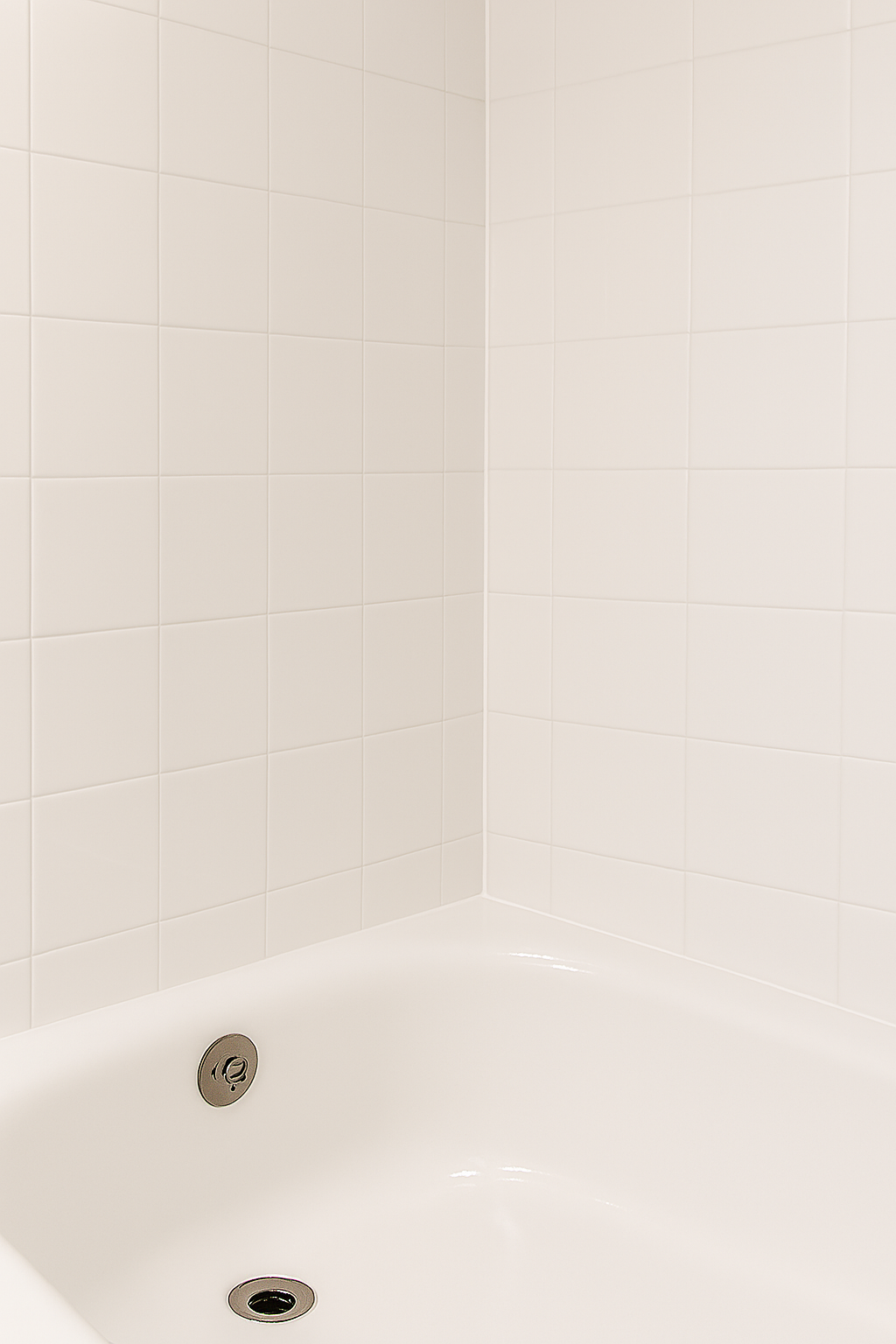 Close-up of a white bathtub with two metal drain covers, surrounded by beige tiled walls.