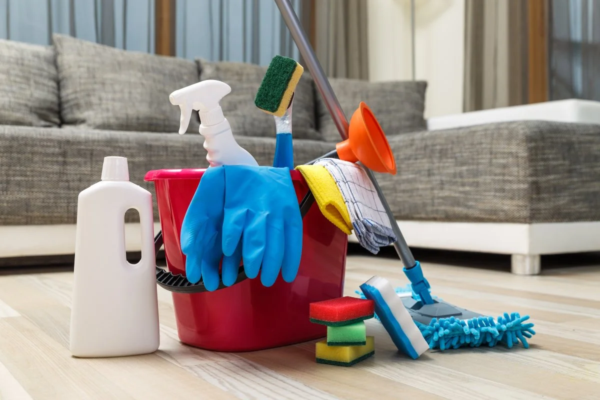 Cleaning supplies including a spray bottle, rubber gloves, sponges, a mop, a scrub brush, a cloth, and other cleaning tools arranged on a wooden floor in front of a gray sofa.