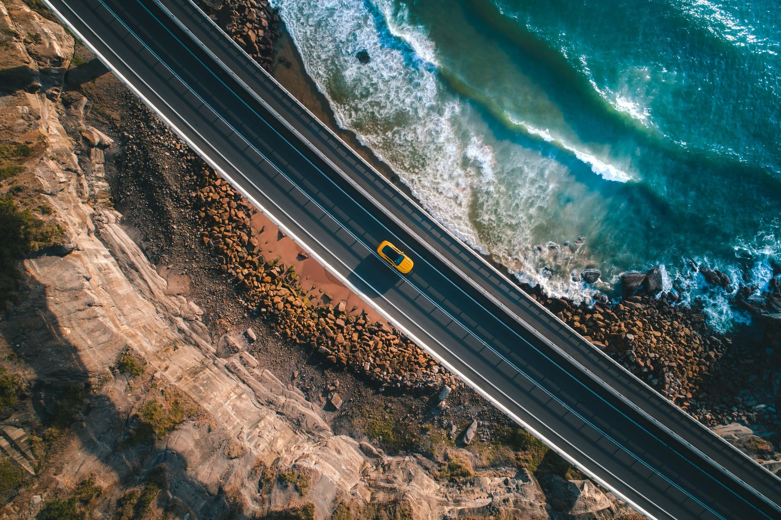 A bird's-eye view of a coastal road with a yellow car driving along it, next to rocky cliffs and ocean waves.