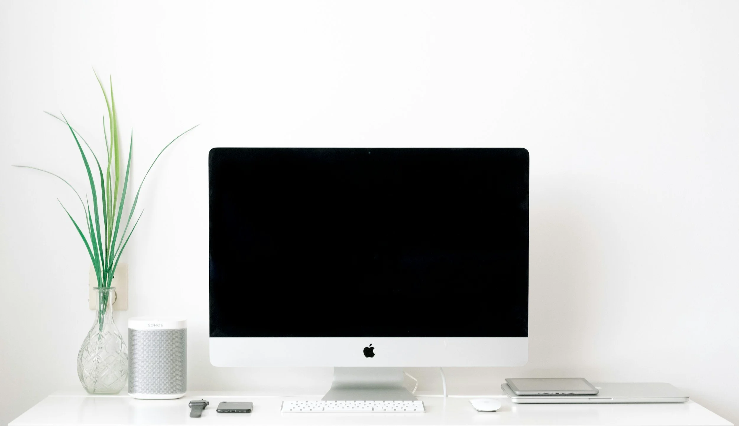 Minimalist workspace featuring an Apple iMac with a black screen, a white keyboard, and a white mouse on a white desk. To the left, a clear glass vase with tall green plant stems, and a small white speaker. To the right, a closed silver laptop and a gray notebook.
