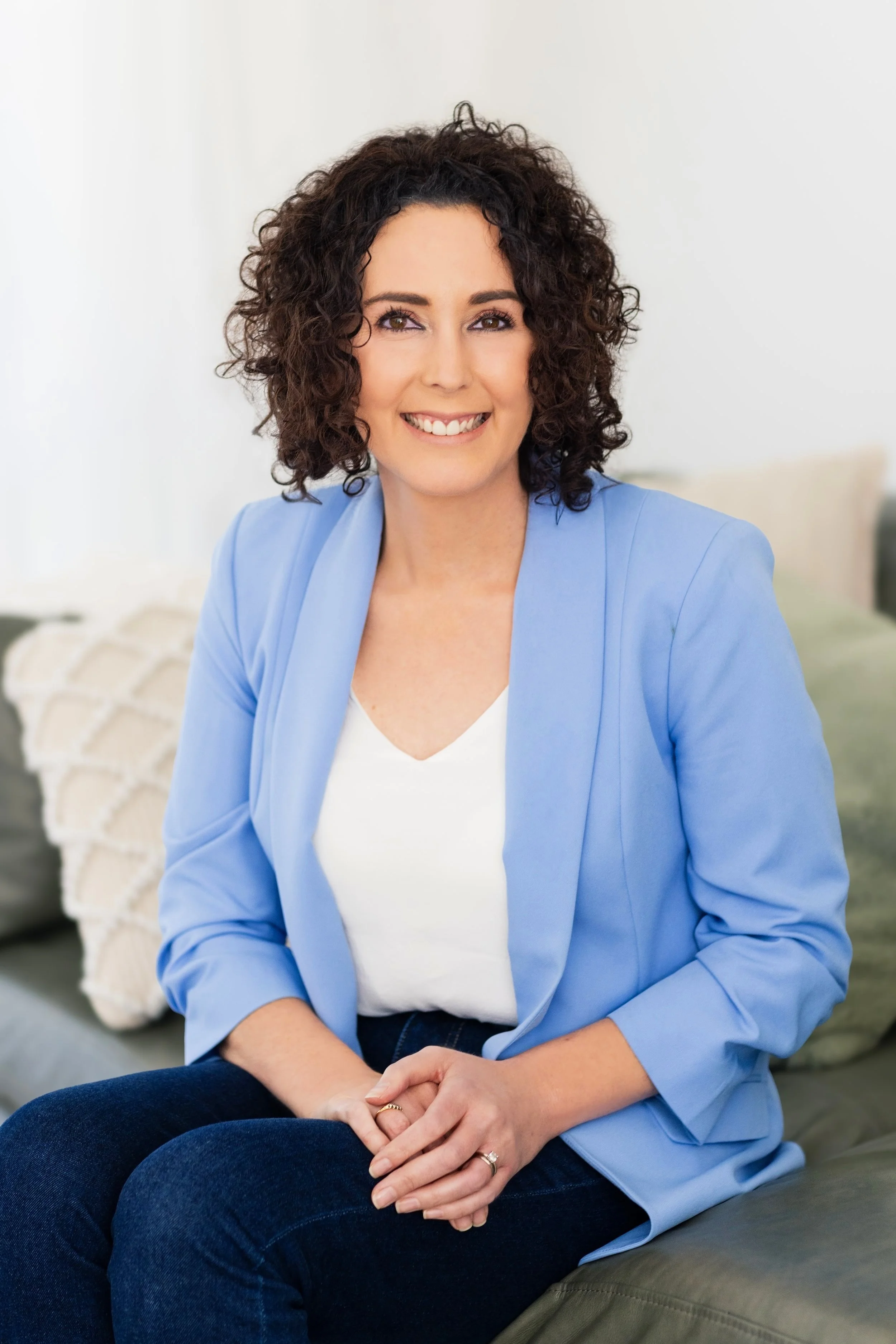 A woman with curly dark hair, wearing a light blue blazer and white top, sitting on a couch in a well-lit room, smiling at the camera.