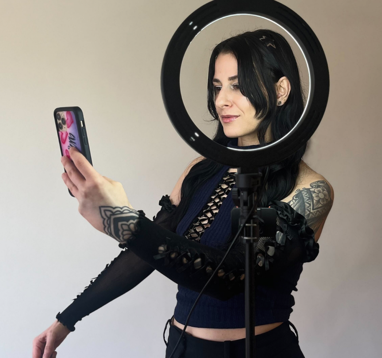 A woman with black hair and tattoos taking a selfie using a smartphone, with a ring light in front of her.
