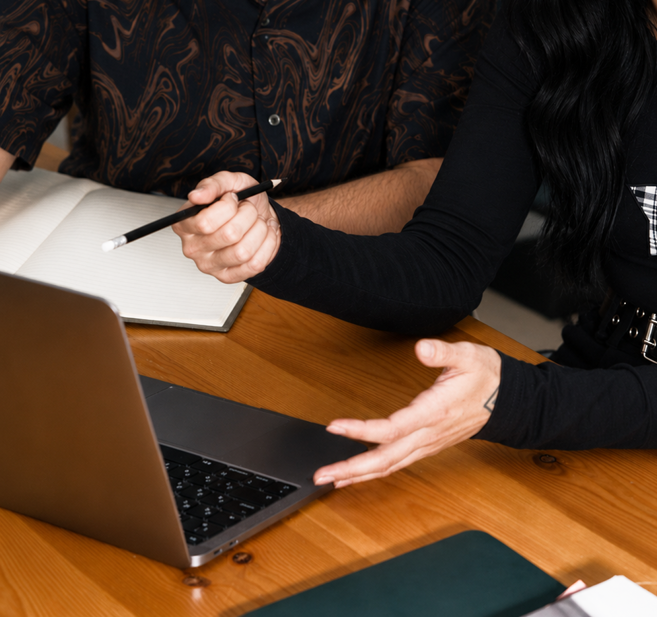 Two people engaging in a discussion at a wooden table, one holding a pen and pointing to a laptop, with an open notebook nearby.