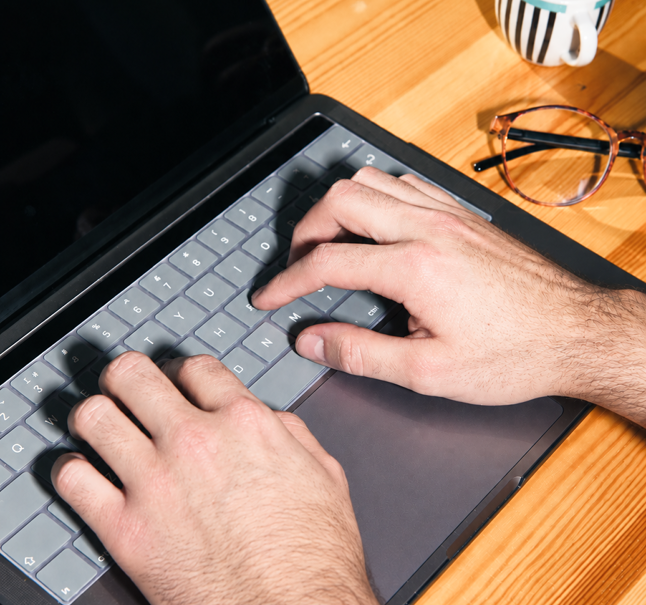 A person typing on a laptop keyboard with a pair of eyeglasses and a striped mug on a wooden table nearby.