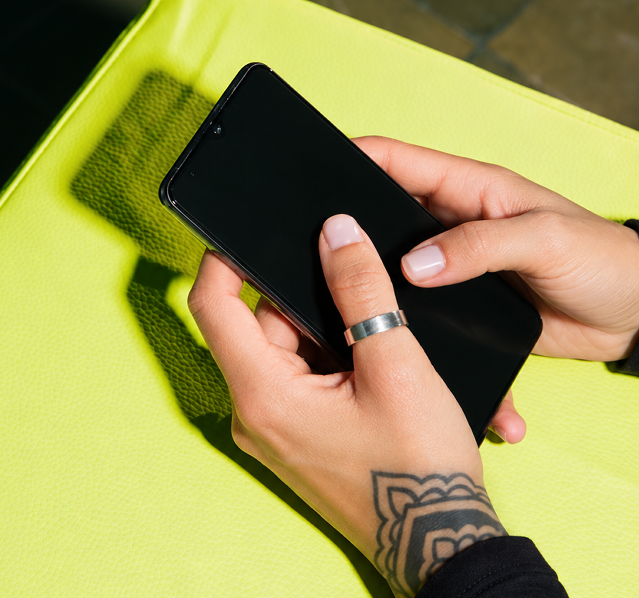Person holding a black smartphone with both hands over a yellow textured surface, with tattoos visible on the wrist and a silver ring on the finger.
