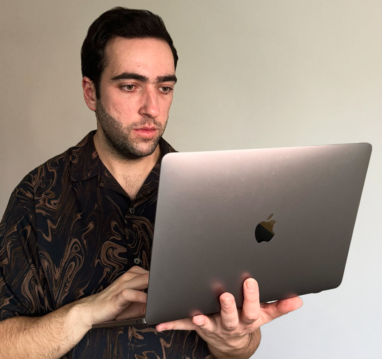 A man with dark hair and beard wearing a black patterned shirt, looking at a silver MacBook laptop.