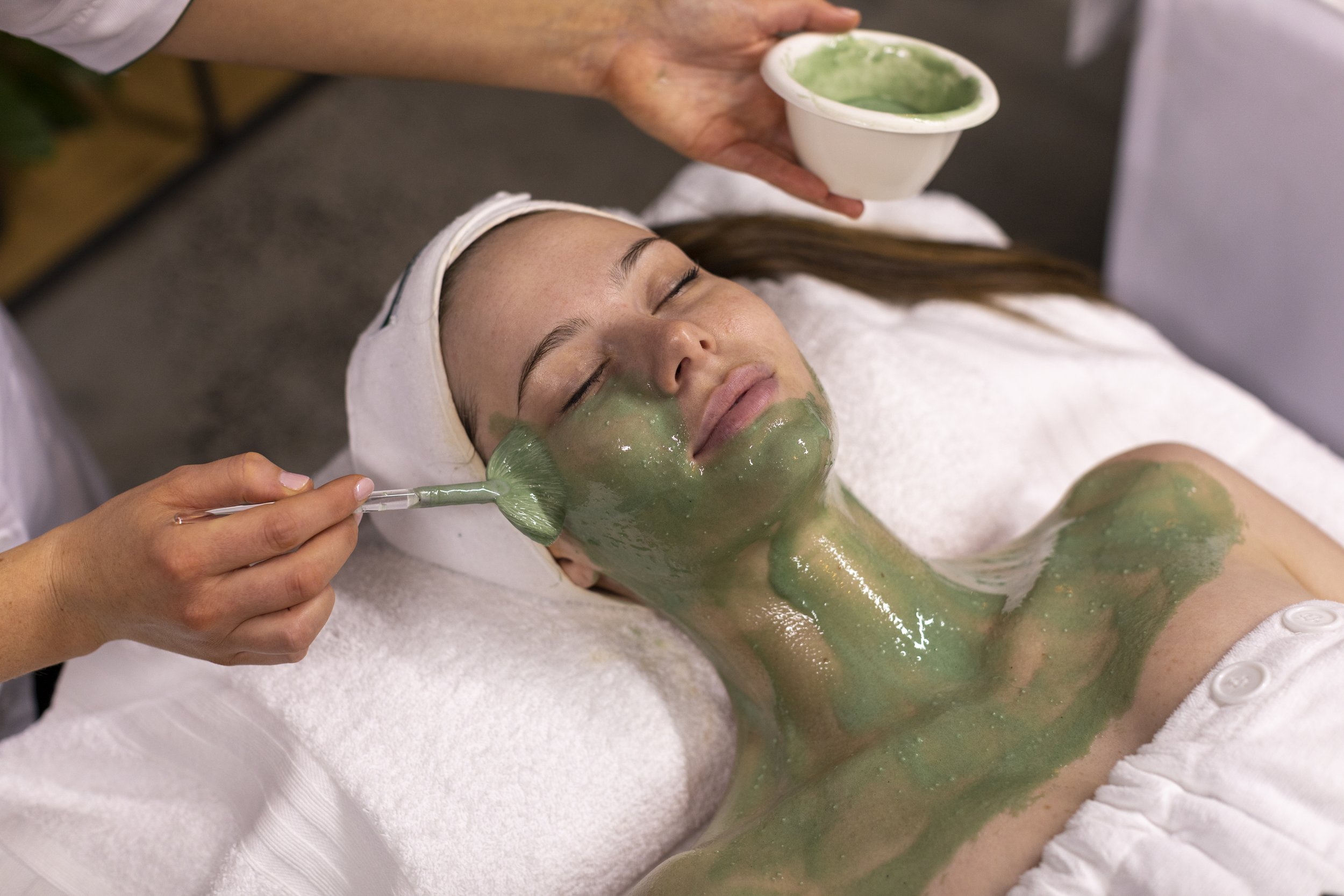 Person receiving a facial treatment with green mask applied to face and neck, lying on a white towel.