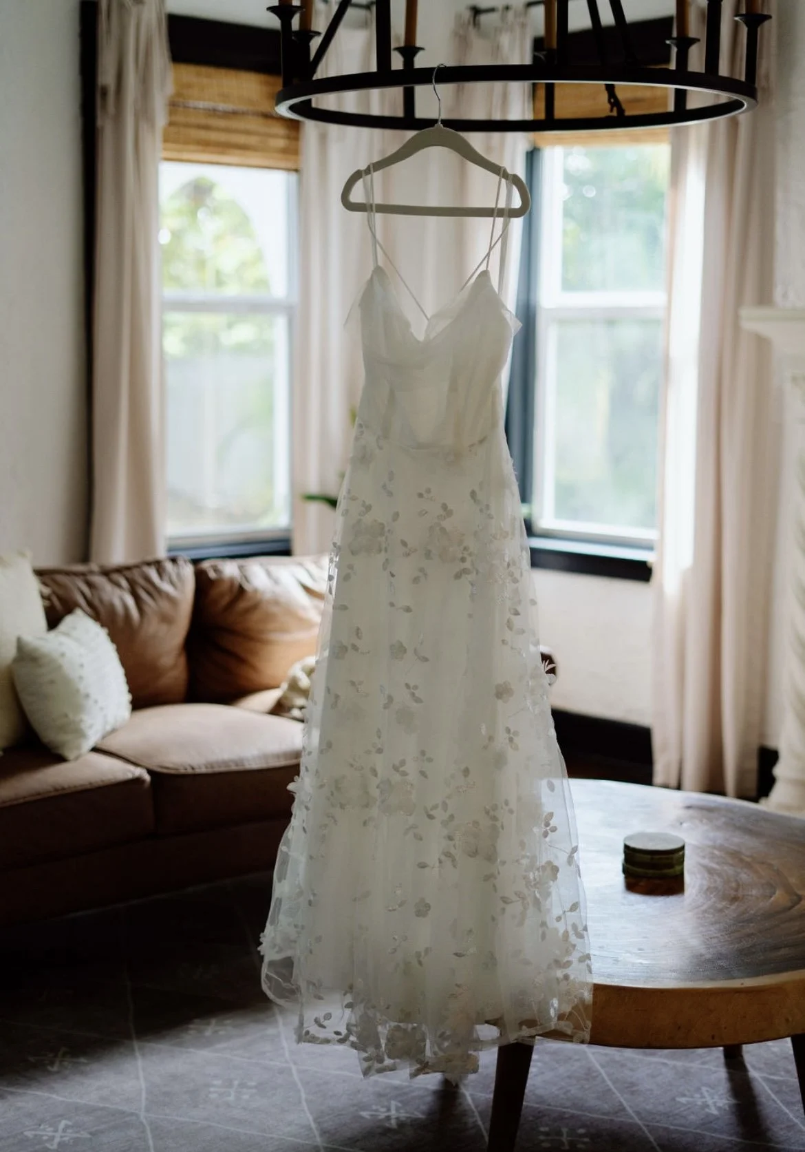 A white wedding dress with floral details hanging from a circular black chandelier in a bright room with windows, a beige curtains, a brown sofa with pillows, and a wooden table.