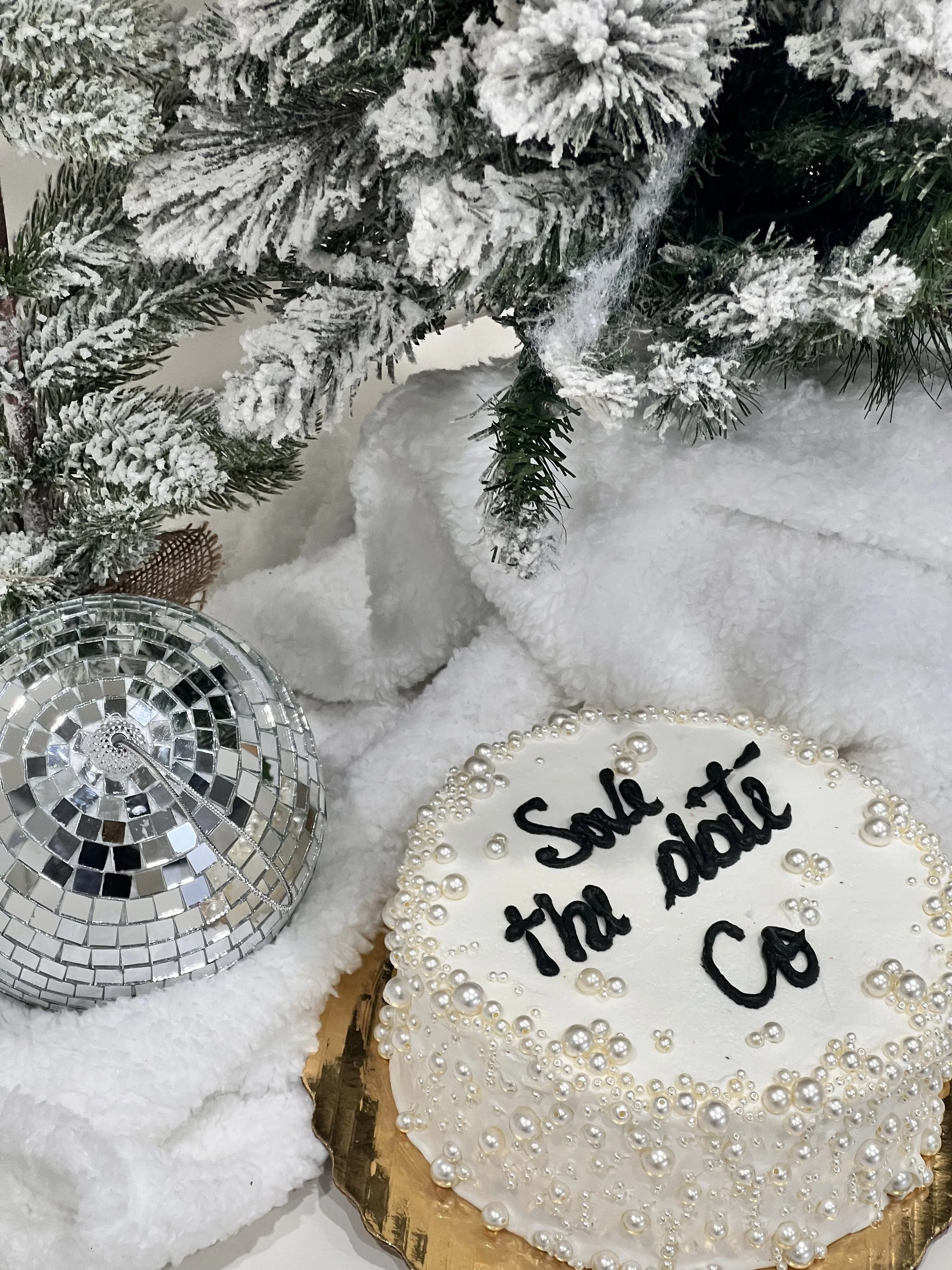 A cake decorated with white icing and pearl-like beads, with the words "sale the date Co" written on top. The cake is placed on a golden base and is surrounded by snow-like fabric, with a part of a snow-covered Christmas tree and a decorative disco ball nearby.