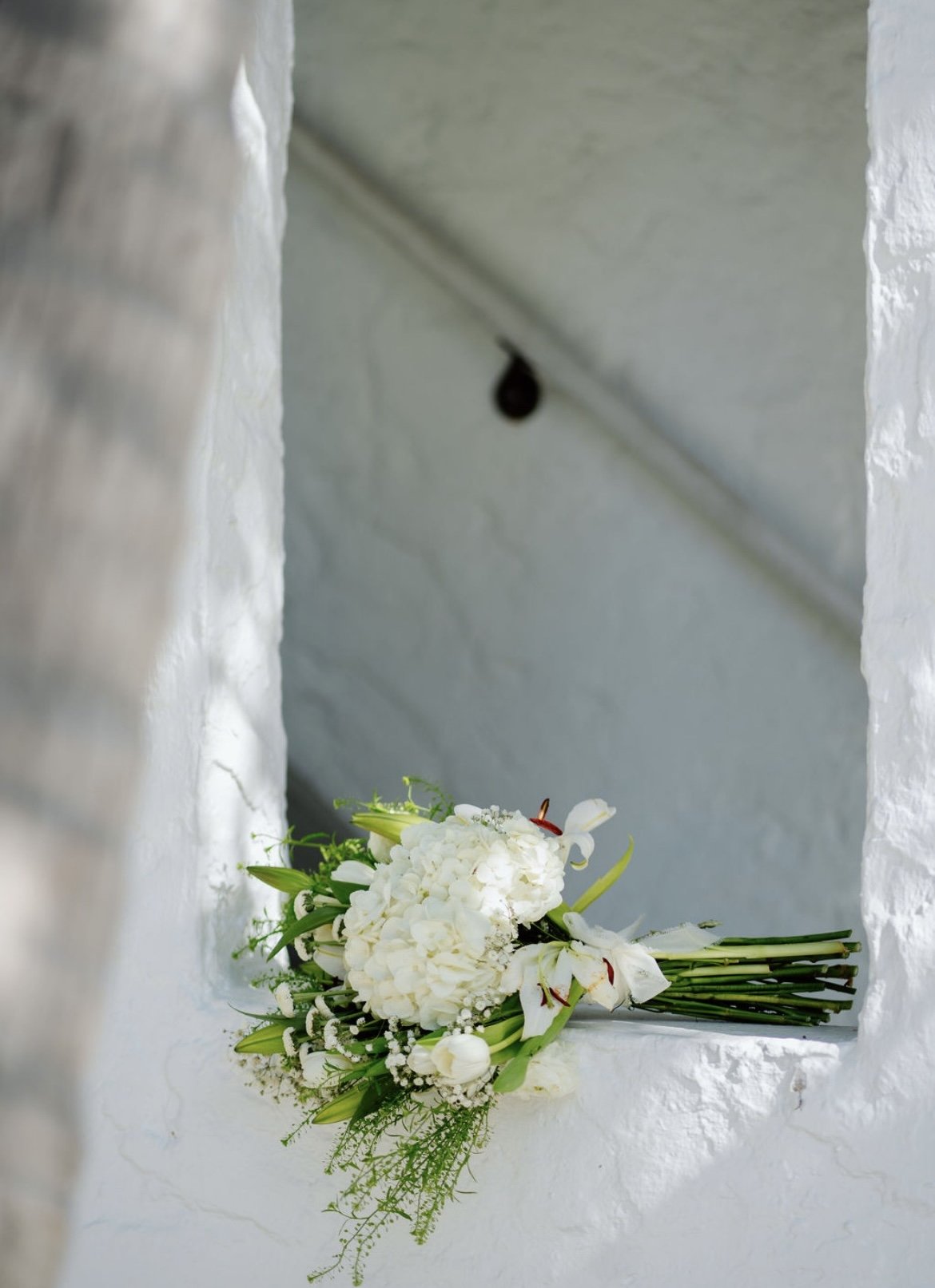 A bouquet of white flowers resting on a white windowsill in front of a white textured wall, with a small black object hanging from the ceiling above.