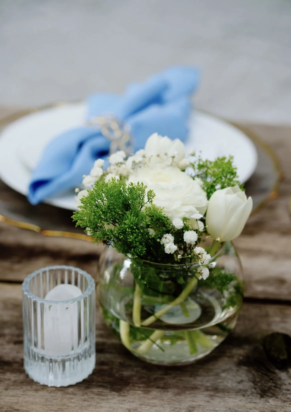A small glass bowl with white flowers and greenery, a white candle in a glass holder, and a blue cloth napkin on a white plate set on rustic wood table.