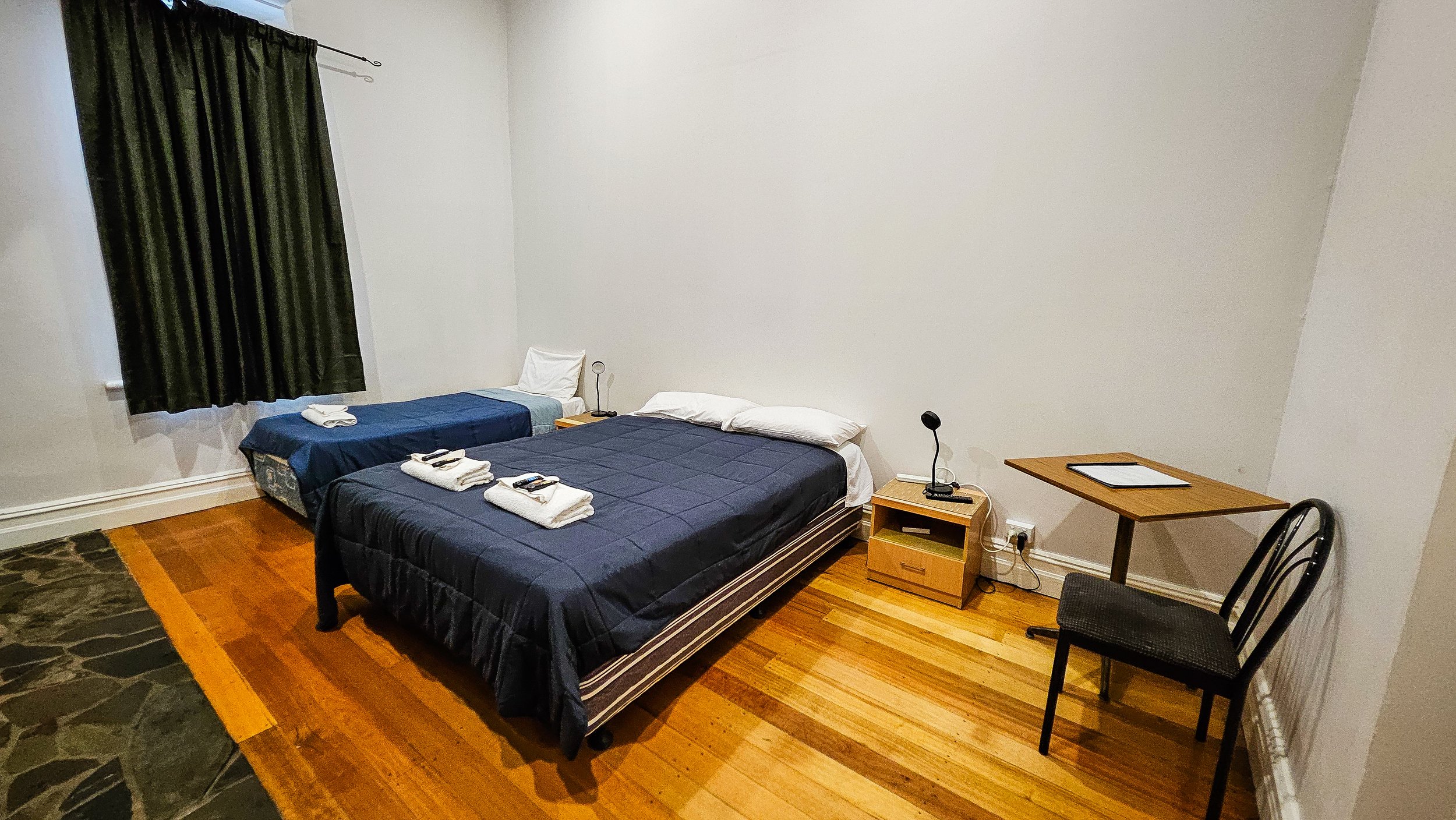 Hotel room with two beds, one with colorful floral bedspread and the other with a multicolored geometric pattern, each with towels folded into swan shapes on top, and a framed picture on the wall.