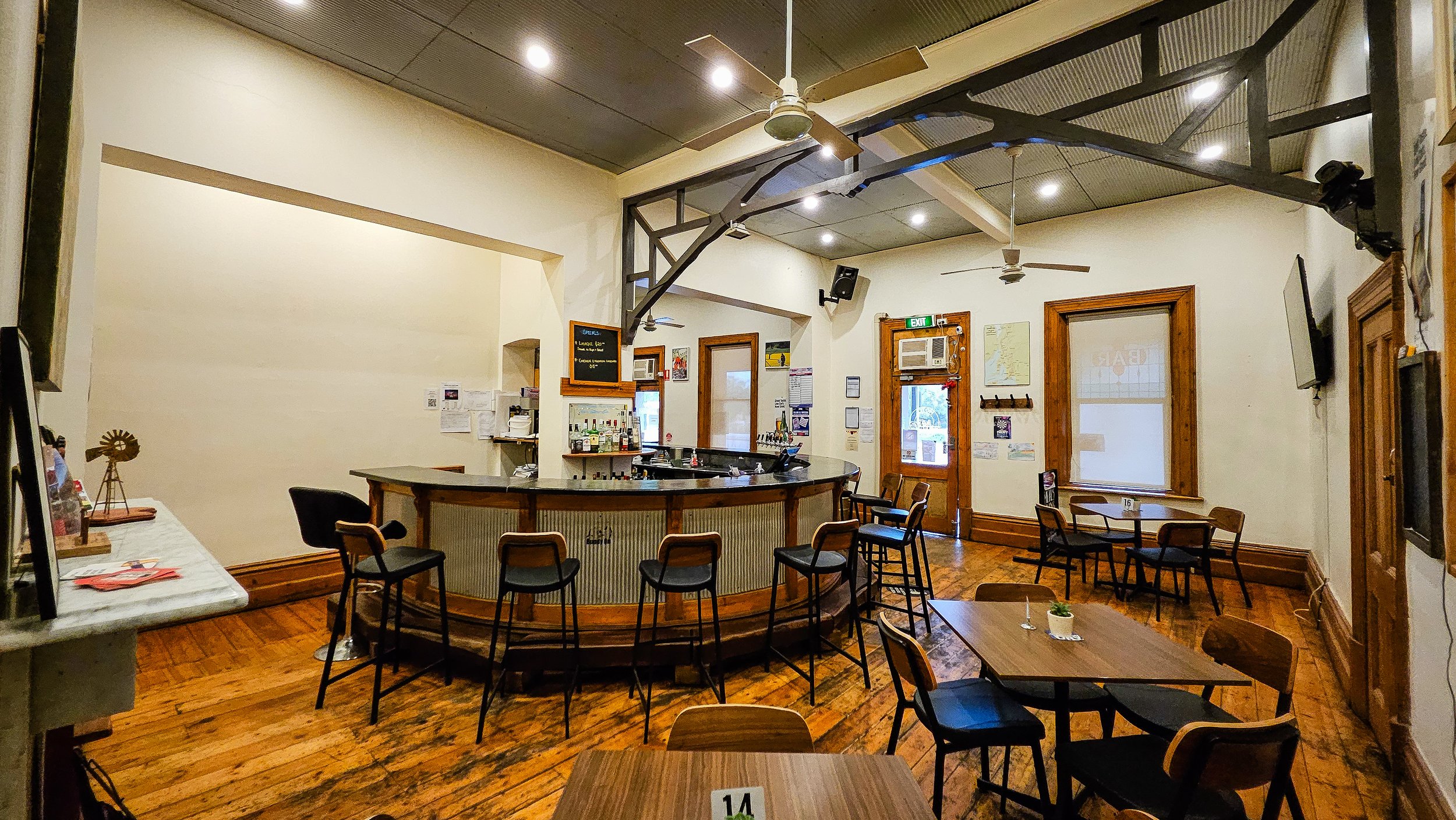 Interior view of a bar with a curved wooden counter, bar stools, and various bottles of liquor on shelves. There are chairs and tables around, a TV on the wall, and a fireplace.