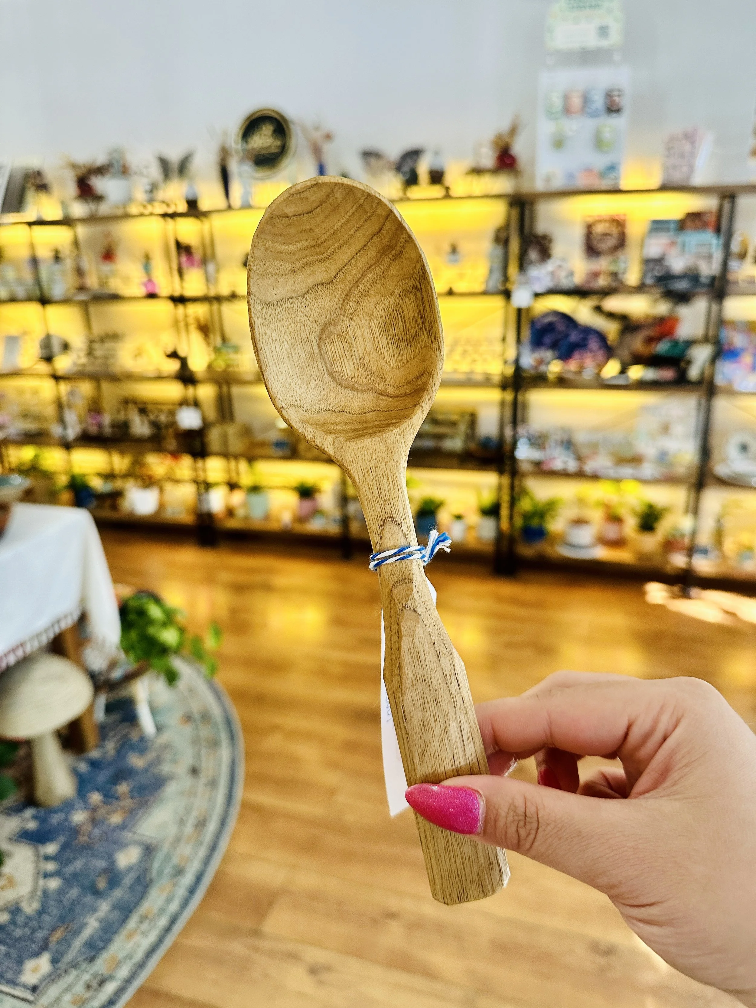 A hand holding a wooden spoon in a store with shelves displaying various items in the background.