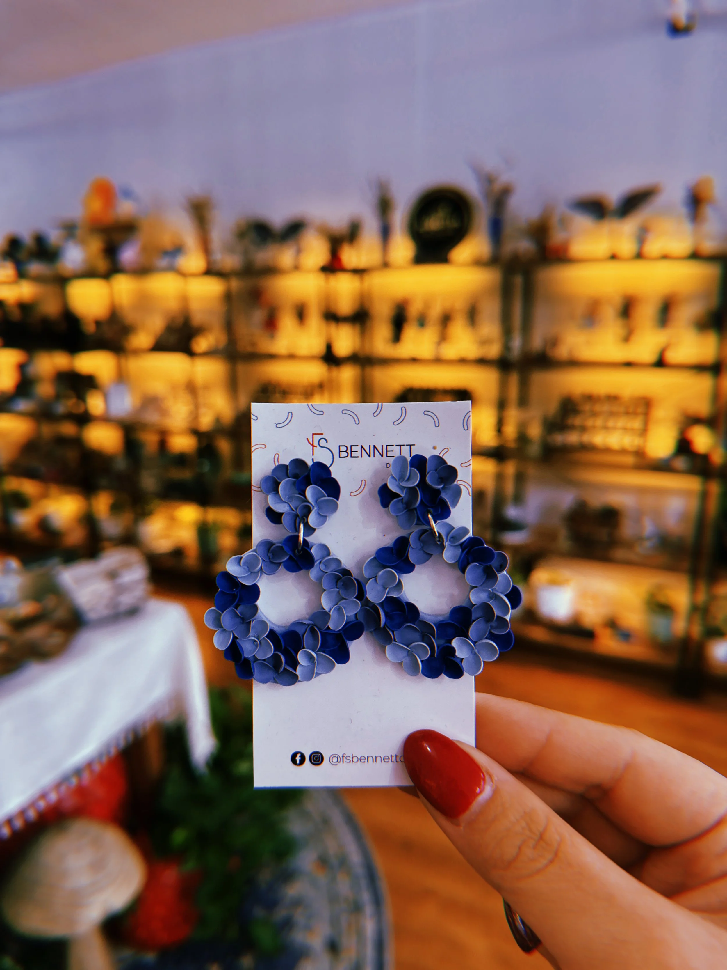 Blue floral-shaped hoop earrings displayed on a jewelry card with brand information, held by a person with red nail polish in a boutique shop with shelves of collectibles and decorative items in the background.