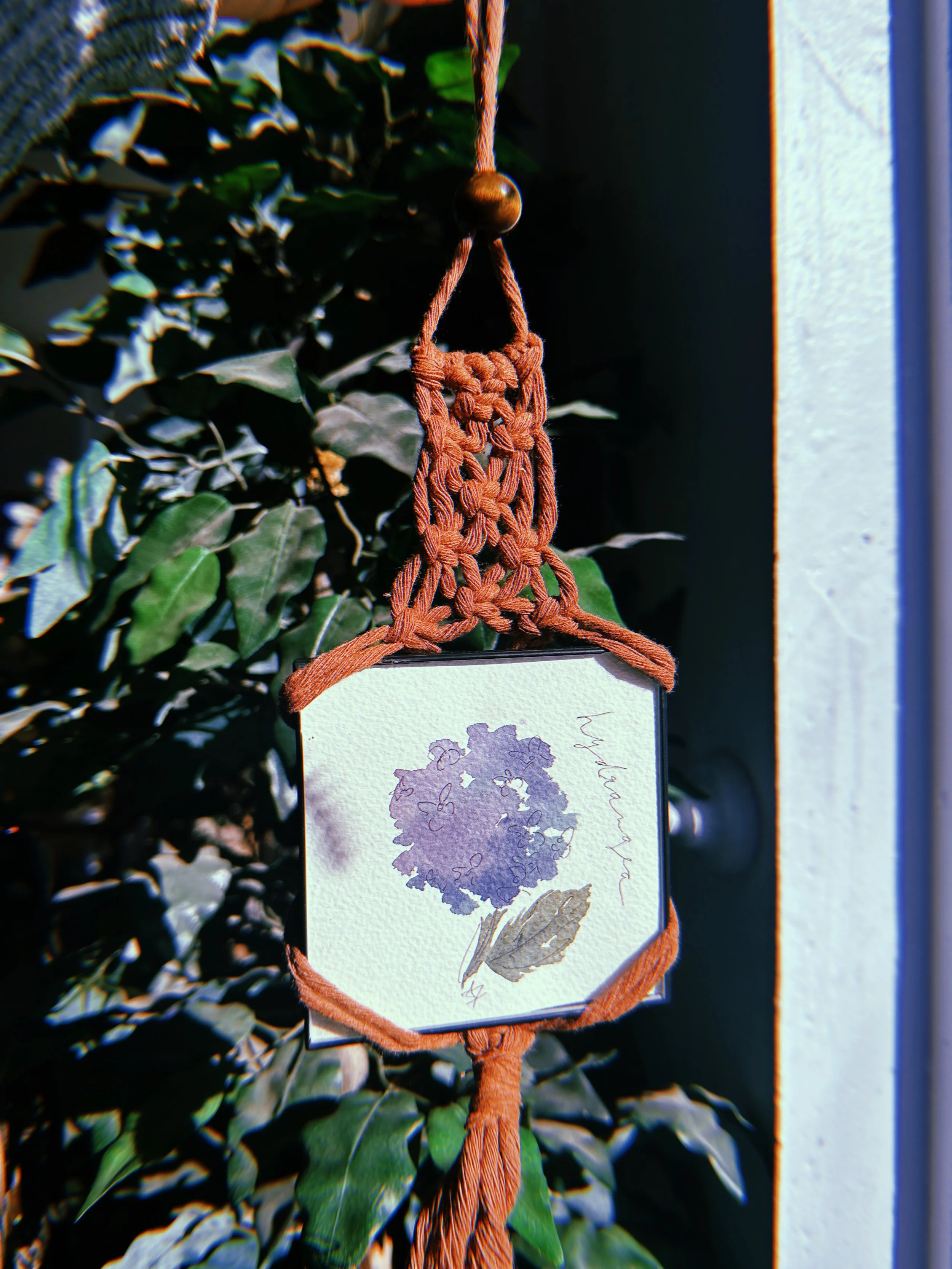 A framed watercolor painting of a purple hydrangea flower with a green leaf, hanging on a plant with green leaves, suspended by a brown macramé holder with a knot at the bottom, attached to a wooden bead at the top.