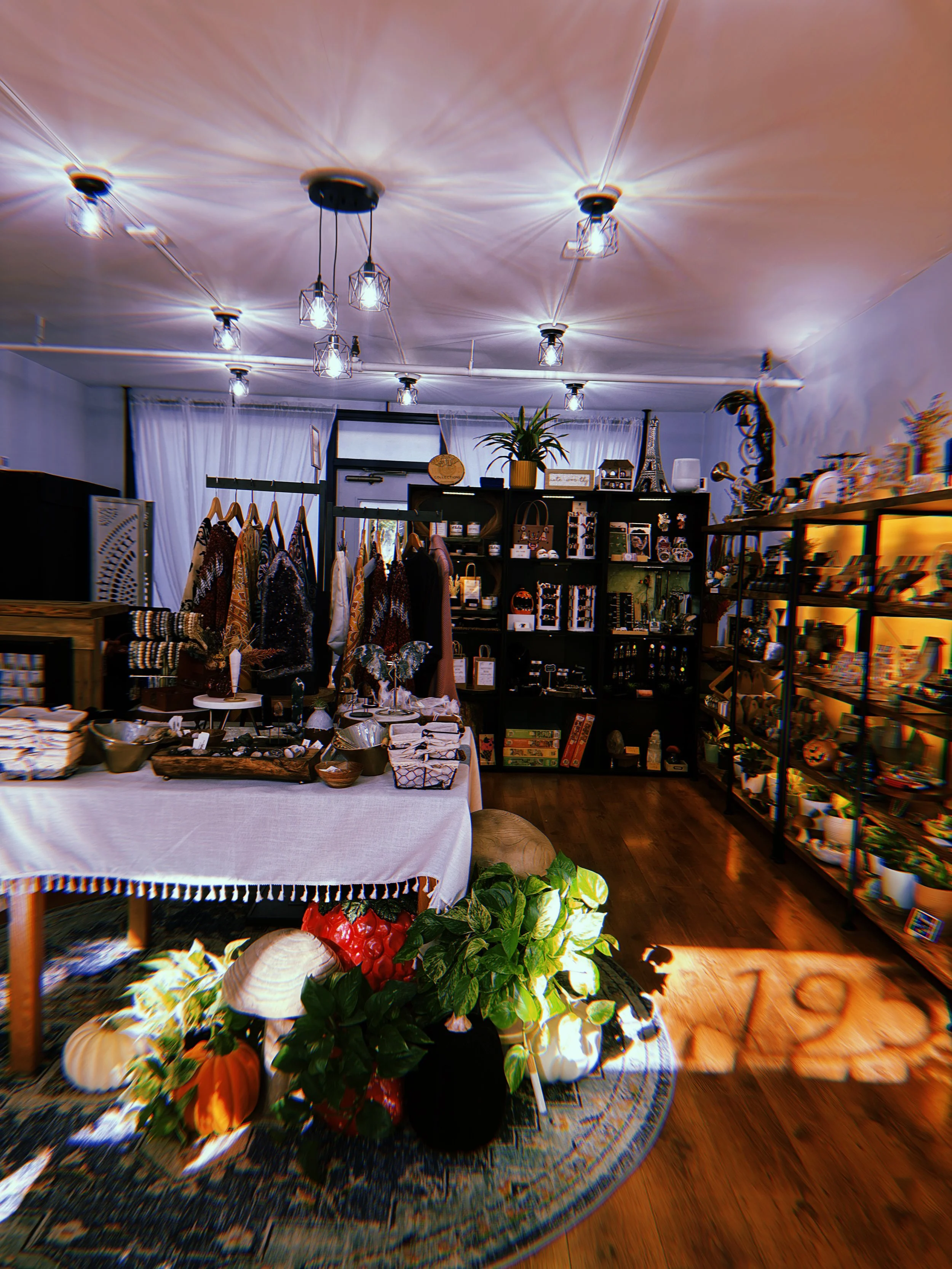 Interior of a small boutique shop with clothing on a rack, decorative items on shelves, and a table displaying accessories and jewelry, with warm lighting and wooden flooring.